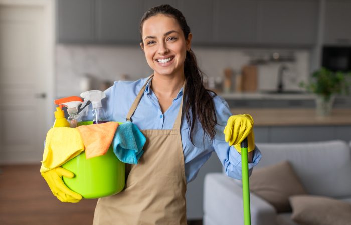 Happy woman equipped with mop and bucket full of cleaning supplies flashes warm smile, ready to tackle household chores with confidence